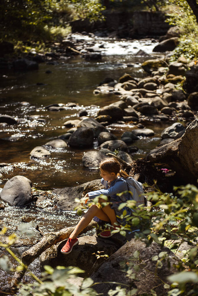 young girl with backpack sitting near rapid river  - Φωτογραφία, εικόνα