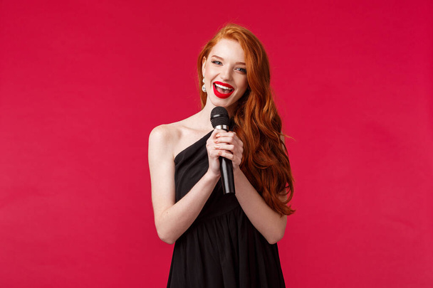 Portrait de femme magnifique jeune femme en robe noire, tenant microphone chantant karaoké ou interpréter des chansons devant le public sur l'événement public, debout fond rouge
 - Photo, image