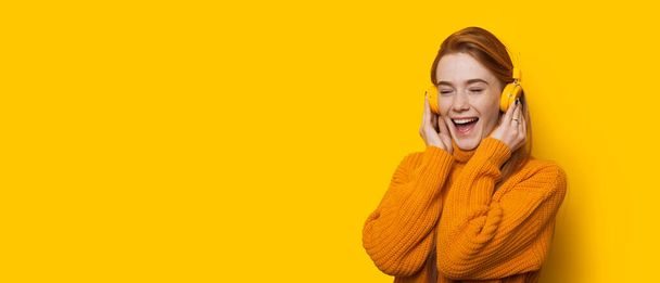 Stunning ginger caucasian lady is listening to music while wearing a sweater on a yellow background with copy space - Zdjęcie, obraz