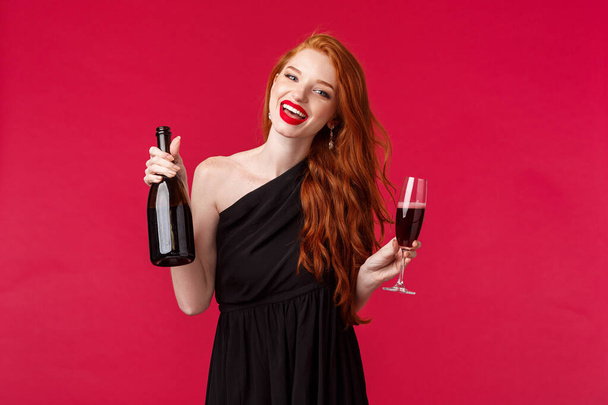 Πορτρέτο της ανέμελης πανέμορφη κοκκινομάλλα γυναίκα πάρτι με φίλους, πίνοντας από το γυαλί, κρατώντας σαμπάνια μπουκάλι και χορό κατά τη διάρκεια bachelorette νύχτα, χαμογελώντας κάμερα φορούν μαύρο φόρεμα - Φωτογραφία, εικόνα