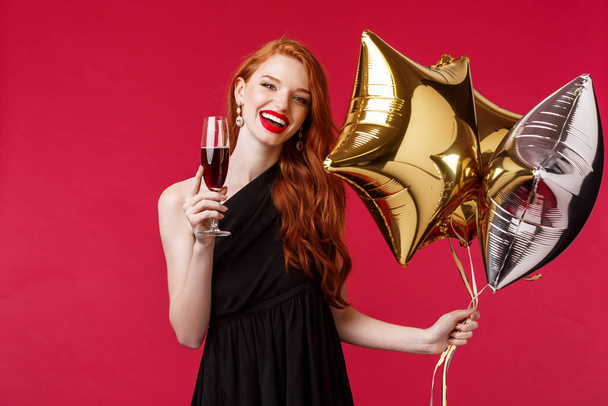 Portrait d'heureuse femme rousse insouciante et magnifique en élégante robe noire faisant la fête, disant toast, acclamations tenant un verre de champagne ou de vin, souriant heureux avec des ballons à la main, fond rouge
 - Photo, image
