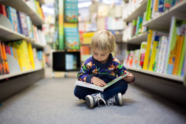 Bir kitapçıda oturup kitap okuyan sevimli küçük bir çocuk. - Fotoğraf, Görsel