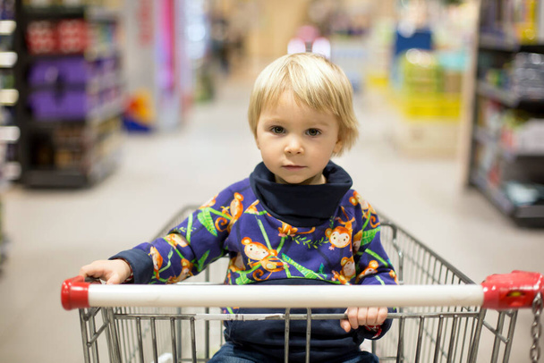 Sweet toddler boy, sitting in shopping cart, mother byuing groceries, people shopping hectically for crysis - Foto, Bild