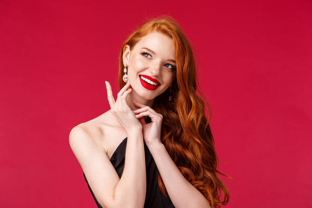 Beauty, fashion and luxury concept. Close-up portrait of gorgeous feminine redhead woman in red lipstick and black dress, touching earring look away with dreamy happy smile - Photo, Image