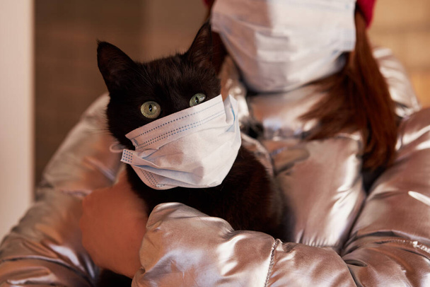 A young woman in a protective mask holds a masked cat in her arms. Conscious citizen in the fight against the epidemic of coronavirus - Fotó, kép