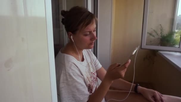 young, pretty girl listens music from smartphone, headphones, on balcony, windowsill, - Footage, Video