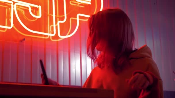 Trendy cute girl blogger in sunglasses taking selfie photo or video using smartphone. Attractive brunette young woman looks at herself with mobile phone and touching her short hair. Red neon light. - 映像、動画