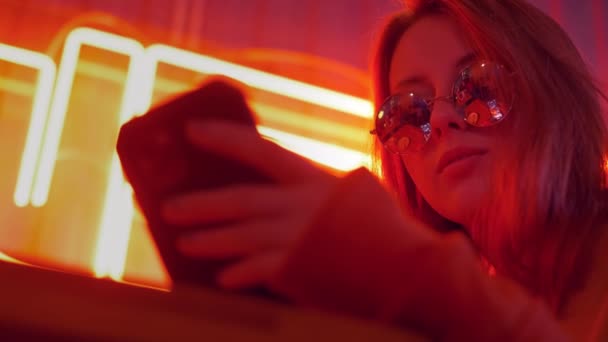 Portrait of attractive smiling girl using online social media and listening music. Cute young woman typing message on smartphone and smile. Dressed casual street style. Colorful red neon lights. - Séquence, vidéo
