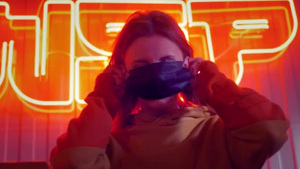 Young beautiful fashionable girl puts on a black medical mask. In a bar or club on the background of neon lights. Protects against epidemics of coronavirus. Pandemic on the planet. - Imágenes, Vídeo