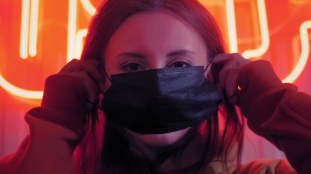 Young beautiful fashionable girl puts on a black medical mask. In a bar or club on the background of neon lights. Protects against epidemics of coronavirus. Pandemic on the planet. - Video