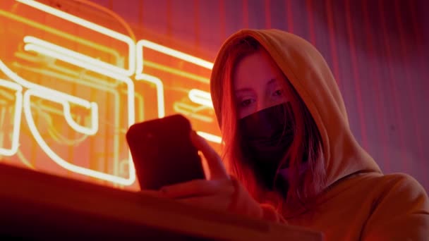 Young beautiful fashionable girl uses a smartphone in a black medical mask. In a bar or club on background of neon lights. Protects against the epidemic of Chinese coronavirus. Pandemic on the planet - Video