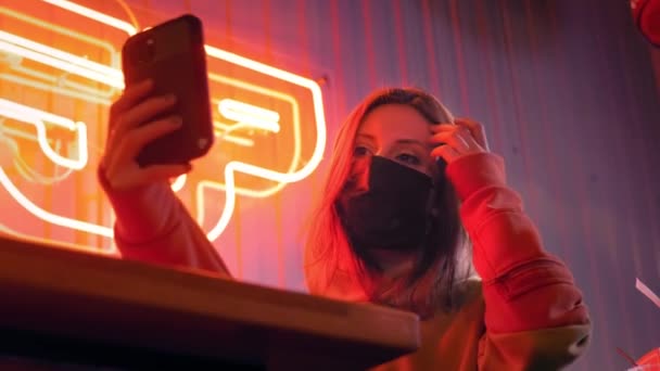 Young beautiful fashionable girl takes a selfie in black medical mask. In a bar or club on the background of neon lights. Protects against the epidemic of Chinese coronavirus. Pandemic on the planet - Кадри, відео