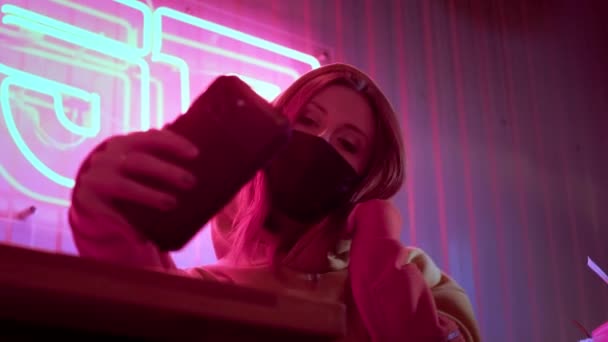 Young beautiful fashionable girl takes a selfie in black medical mask. In a bar or club on the background of neon lights. Protects against the epidemic of Chinese coronavirus. Pandemic on the planet - Кадри, відео