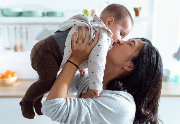 Shot of happy young mother lifting her cute daughter up high in air while kissing in the kitchen at home. - Photo, Image
