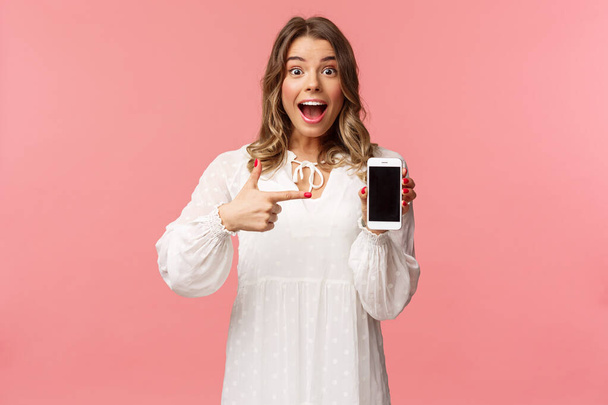 Portrait of impressed, excited young blond woman showing something awesome on display, pointing mobile phone screen and smiling astonished, brag with her recent match on dating app - Photo, image