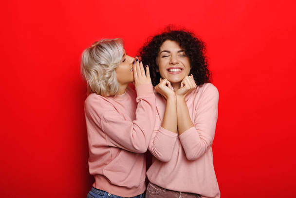 Blonde haired caucasian woman whispering something to her curly haired friend on a red background - Fotó, kép