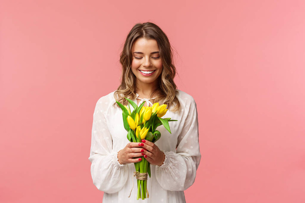 Holidays, beauty and spring concept. Portrait of lovely, romantic blonde girl in white dress, holding yellow tulips, looking at beautiful flowers, smiling and blushing, pink background - Foto, imagen