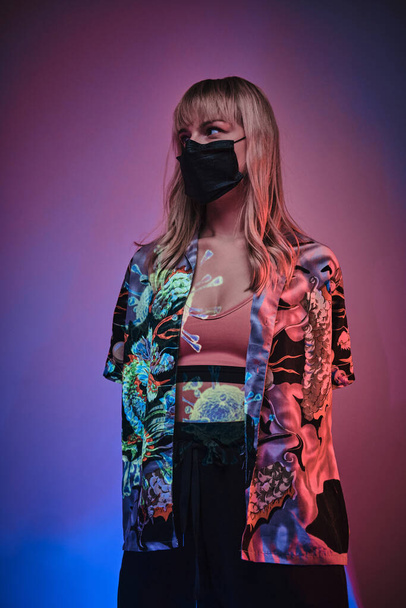 Junge erwachsene Frau posiert in hellem Neon-Studio mit medizinischer Schutzmaske - Foto, Bild