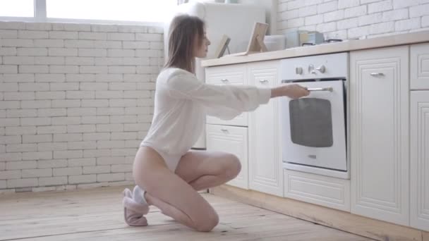 Side view portrait of sensual young woman looking inside the oven in kitchen. Brunette Caucasian girl in white body using kitchen utility in the morning. Lifestyle, beauty, foods. - Materiał filmowy, wideo