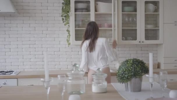 Joyful young Caucasian woman coming into modern kitchen, taking rolling pin and singing into it. Positive brunette girl having fun at home. Lifestyle, joy, happiness, leisure. - Video, Çekim