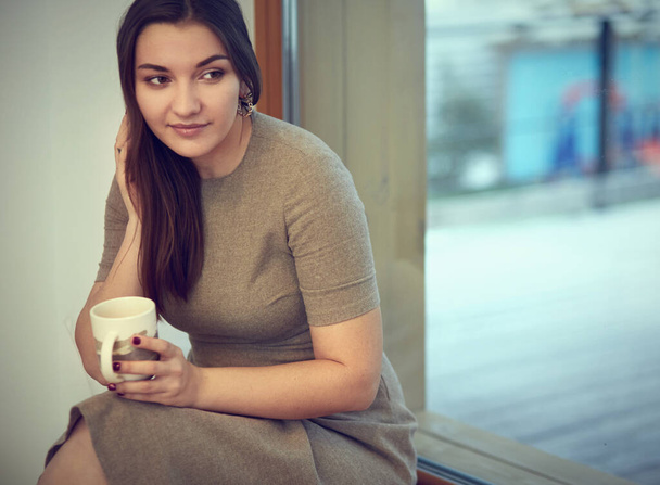 Young and cute lady sitting on the windowsill and looking out the window with cup of coffee - Photo, image