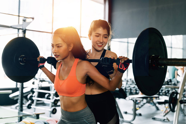 Asian personal trainer coaching a bodybuilding woman to perform the exercise squat with barbell in the fitness gym. - 写真・画像