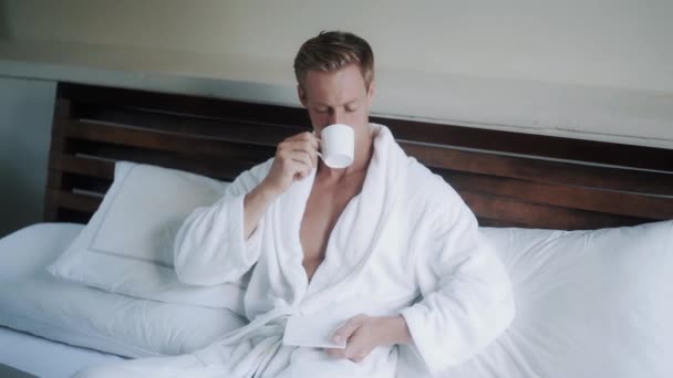 young man in bathrobe drinks coffee in bed near laptop - Кадры, видео