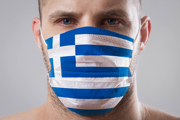 Νεαρός με πονεμένα μάτια σε ιατρική μάσκα ζωγραφισμένος στα χρώματα της εθνικής σημαίας της Ελλάδας - Φωτογραφία, εικόνα