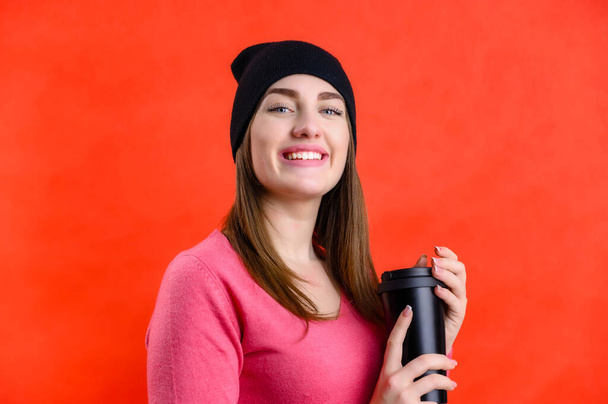 Portrait of a pretty Caucasian woman with a street cup of coffee in a black hat in a pink sweater on a red background. Model smiles, shows joyful emotions - Fotoğraf, Görsel