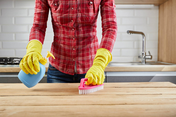 Woman in yellow protective glove with blue plastic bottle and brush, wooden table, kitchen background - 写真・画像