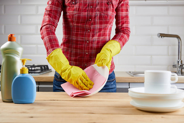 Woman in yellow protective glove with plates, dishes, plastic bottles and cloth, wooden table, kitchen background. - Foto, Imagem