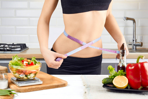 Young sports girl with naked torso measuring her waist with measuring tape in the kitchen. Ripe tomatoes, peppers and salad on the table. diet and weigh loss concept - Fotografie, Obrázek