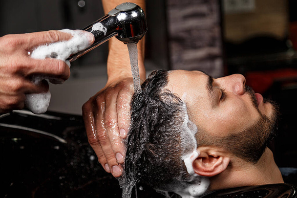 mladý tmavovlasý muž indického nebo asijského vzezření v holičství na černém křesle. holič si umývá hlavu šamponem. - Fotografie, Obrázek