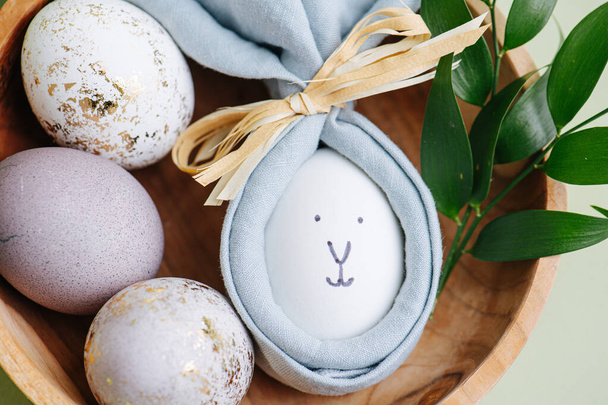 Easter holiday concept. Closeup of handmade rabbit made of egg and napkin on a plate decorated with leaves and painted eggs - Фото, изображение