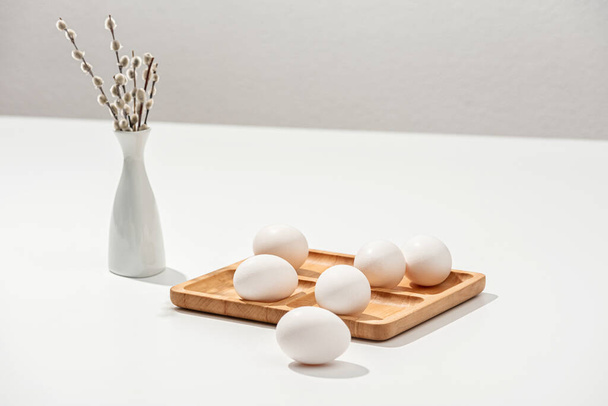 Naturaleza muerta de Pascua. Huevos de Pascua en un soporte de madera y un jarrón blanco con ramitas de sauce en una superficie blanca
. - Foto, Imagen
