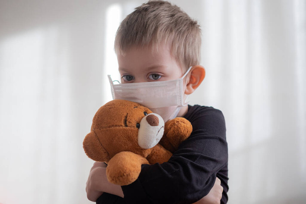 テディベアを抱きしめる医療マスクの男の子。家庭用隔離コロナウイルス｜covid-2019隔離 - 写真・画像