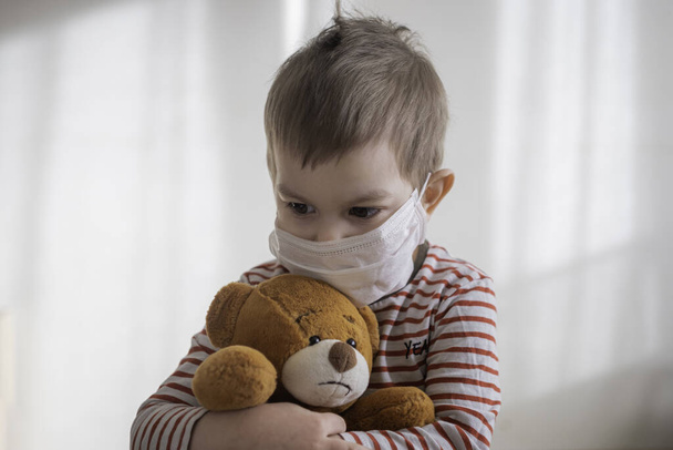 Toddler boy in medical mask embracing his teddy bear. Home isolation coronavirus covid-2019 quarantine - Photo, Image
