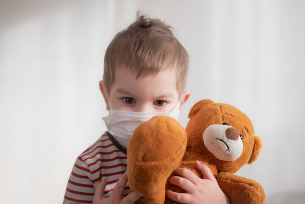 Toddler boy in medical mask embracing his teddy bear. Home isolation coronavirus covid-2019 quarantine - Photo, Image