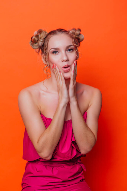 The girl in a red dress on a orange background in the studio. Blonde girl with two hair knots holding her chicks with hands showing astonishment - Photo, Image