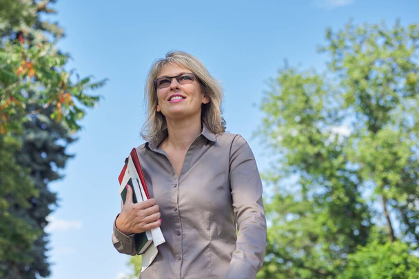 Business confident mature woman walking with documents outdoor. Beautiful blonde female in glasses, gray shirt, sky park background - Φωτογραφία, εικόνα