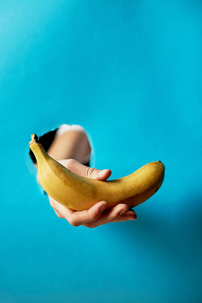 A hole in the paper, a hole. Hand holding a banana, on a blue background.Vegetarianism, the diet. Copy space, trend.Background the city of - Фото, зображення