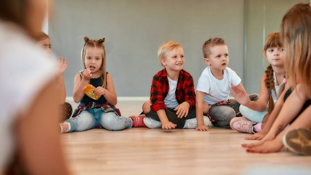 Little dancers. A group of little cute girls and boys in casual clothes talking to each other while sitting in circle during choreography class in the dance studio - Φωτογραφία, εικόνα