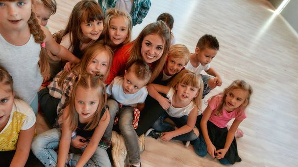 一緒にポーズ。ポジティブでかわいい子供たちと幸せな女性のダンストレーナーは、ダンススタジオで床に座っている間にカメラと笑顔を見て - 写真・画像