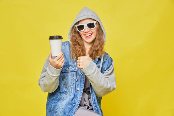 Статильна кавказька дівчина в джинсах і окулярах 8 біт на жовтому тлі. Зберігати одноразову каву. Копіюю простір. Зачиніть.. - Фото, зображення