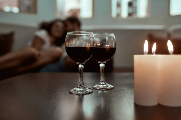 Romance hold a relationship tight. Wine glasses and candles on table with couple relaxing in the background on couch - Foto, afbeelding