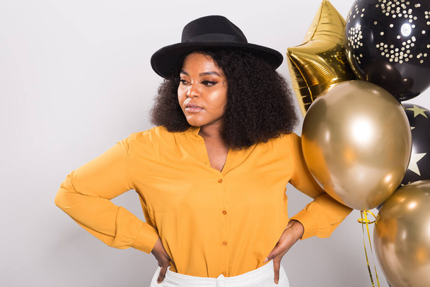 Праздники, вечеринки и веселая концепция - портрет улыбающейся молодой афро-американской девушки, выглядящей стильно на белом фоне с воздушными шарами
. - Фото, изображение