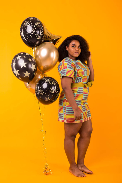 Vacanze, festa e divertimento - Ritratto di giovane donna afro-americana sorridente che sembra dolce su sfondo giallo con palloncini in mano
. - Foto, immagini