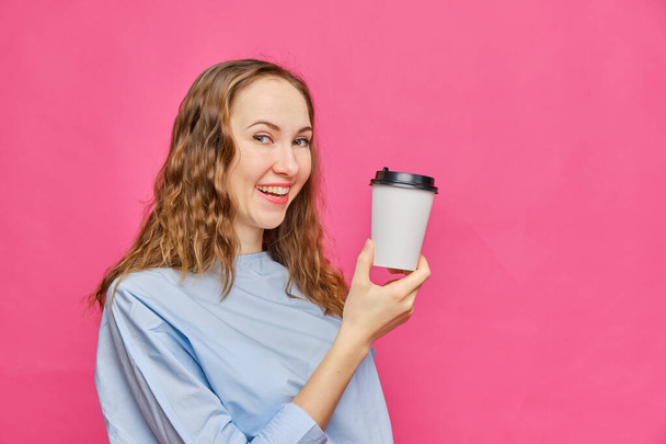 Stylowa biała dziewczyna w jasnoniebieskim t-shircie pokazująca kawę rozpuszczalną w papierowym kubku na różowym tle. Odbiór. Zamknij się.. - Zdjęcie, obraz