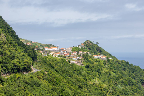 Dramatische Szene an der Nordküste Madeiras mit dunklen Wolken, hohen Bergen und einer Küstenlinie mit steilen Klippen. in den vorderen grünen Feldern und dem kleinen Dorf São Jorge. Blaues Meer mit großen Wellen. - Foto, Bild