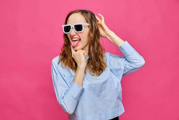 Stylish caucasian girl in a pale blue t-shirt and 8-bit glasses shows tongue and grimaces on a pink background. Close up. - Photo, Image